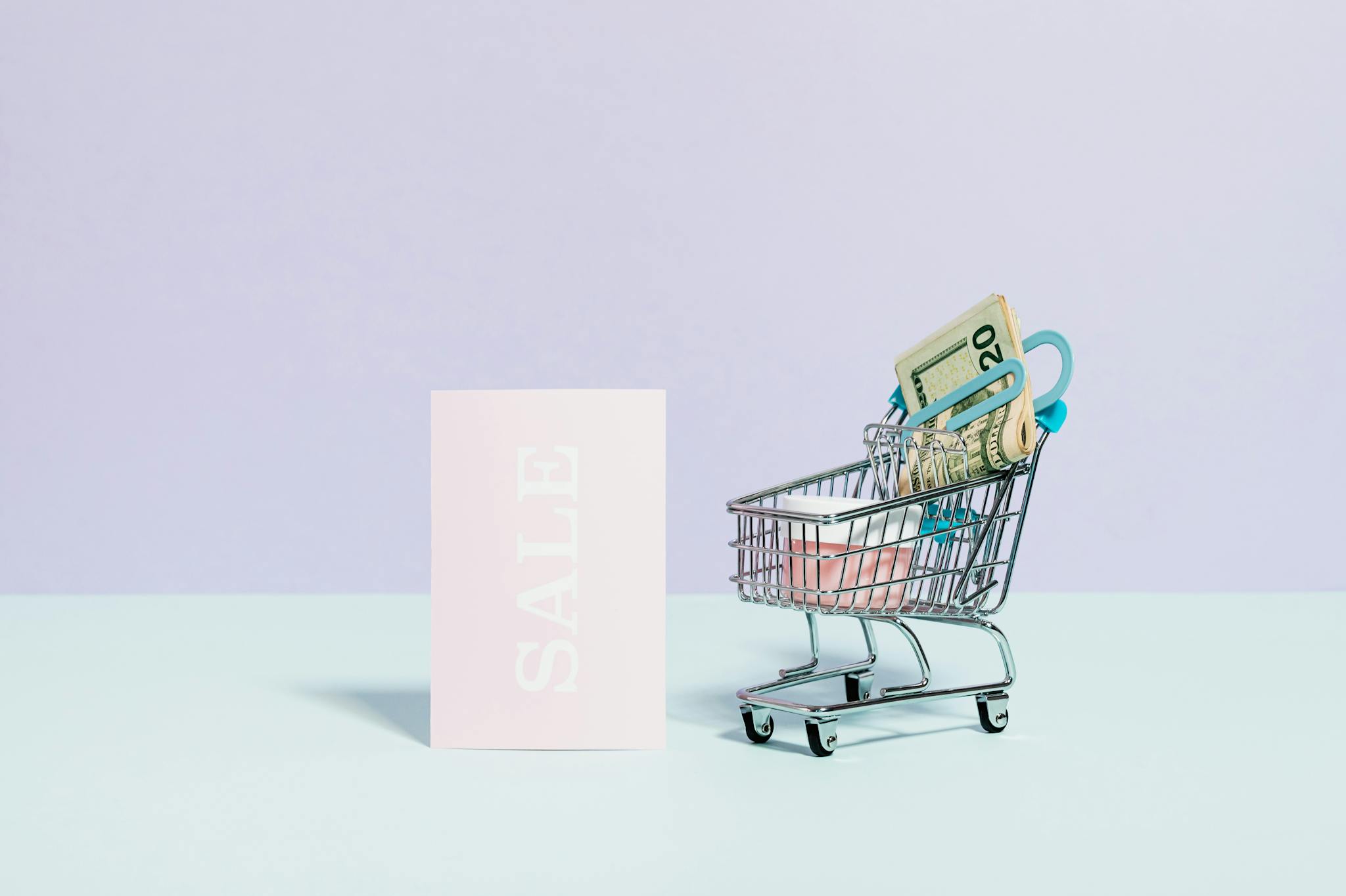 Mini shopping cart with cash alongside a sale sign on a pastel background, perfect for online shopping themes.